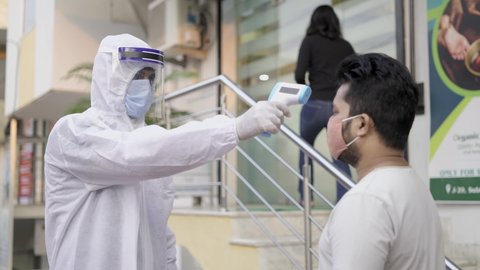 Indian security personnel wearing a disposal personal protection kit with protective face shield and face mask checking temperature of a male client by using a thermometer outside a corporate office.