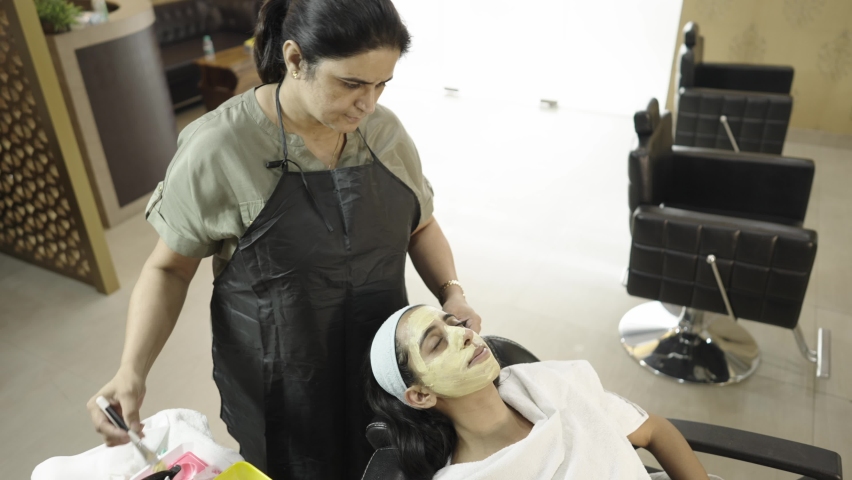 A long shot of a lady beautician in apron giving a skin treatment by applying face mask cream using a brush on the face of a young Indian Asian female sitting in a comfy chair in a beauty parlor Royalty-Free Stock Footage #1066161376