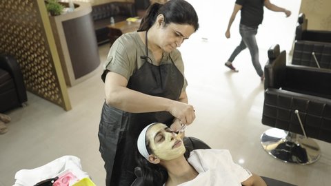 A long shot of a lady beautician in apron giving a skin treatment by applying face mask cream using a brush on the face of a young Indian Asian female sitting in a comfy chair in a beauty parlor