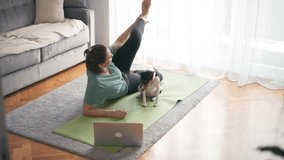 Beautiful young adult woman taking an online yoga class on her laptop and making exercises on the mat in the living room with her cute basenji dog.