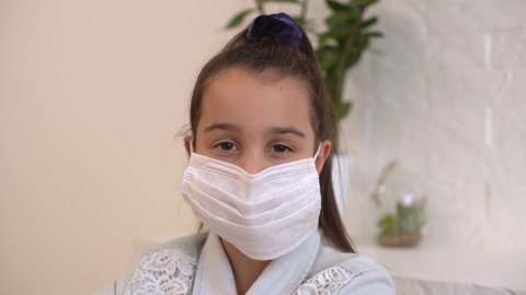 Little girl takes off her protective medical mask and smiles. Stay home mom. Coronavirus or COVID-19. Cute little girl in a protective mask at home in a bright sunny room