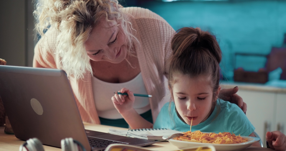 Granddaughter eats spaghetti while doing homework using a laptop, the young grandmother helps the girl to have lunch. Family happy day, multigeneration