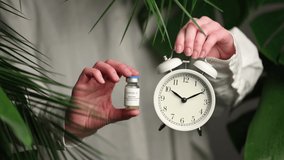 Woman in white shirt show alarm clock and vaccine near palm leaves.