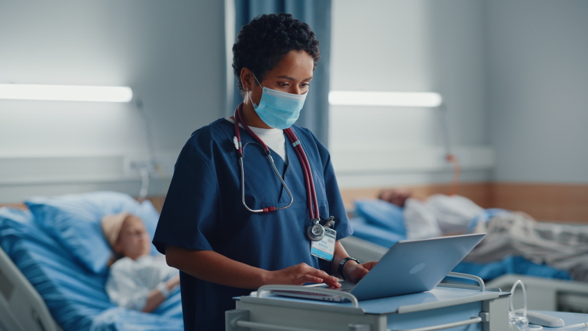 Hospital Ward: Professional Black Latin Female Head Nurse or Doctor Wearing Face Mask Uses Medical Computer. In the Background Patients in Beds Recovering Successfully after Sickness and Surgery Royalty-Free Stock Footage #1066174369