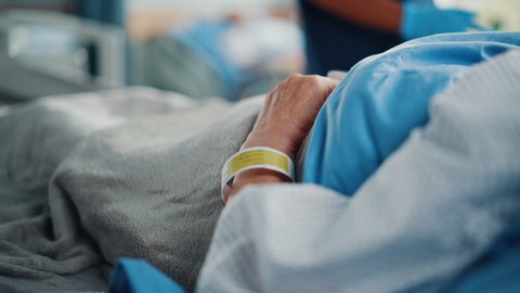 Hospital Ward Bed: Recovering Old Patient Lying in the Hospital Bed Sleeping, Her Fragile Hands Resting on a Blanket, on it Information Wristband. Focus on the Hand. Deeply Emotional Cinematic Shot