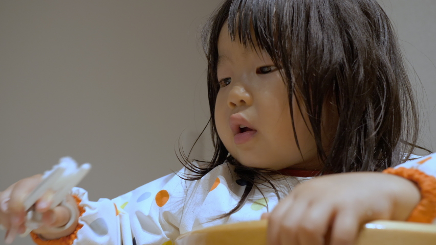 Asian preschool toddler girl who skillfully eats white rice food with orthodontic plastic chopsticks for children at home. Growth and parenting. Kid in polka dot apron sitting wooden child chair