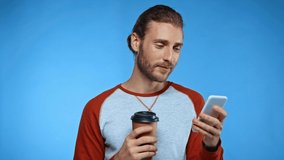 happy man with coffee to go messaging on smartphone isolated on blue