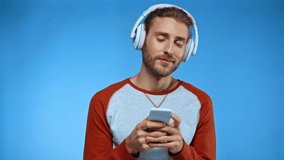 cheerful man in headphones messaging on mobile phone isolated on blue