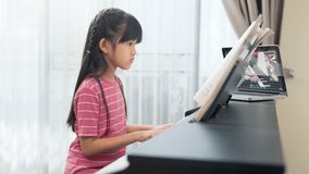 Homeschool Asian little young kid girl learning piano from computer connecting to internet music online class by school teacher. New normal lifestyle and education, student study at home concept.