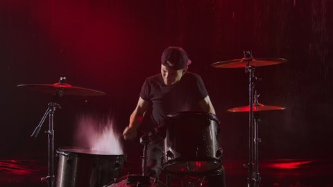 Progressive young man enthusiastically plays the drum kit with drumsticks. Rock musician hits drums in rain in smoky dark studio with red lights. Water splash and splatter rise upwards in slow motion.