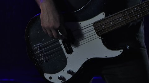 Rock musician plays a black white bass guitar in a dark studio against the background of falling rain drops. Close up of male hands play on strings. Slow motion.
