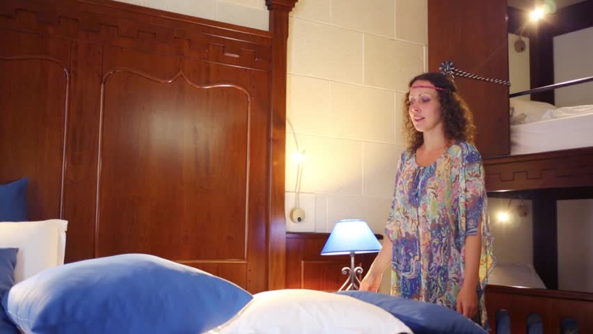 Pretty woman in ethnic dress corrects pillows on double bed in hotel room