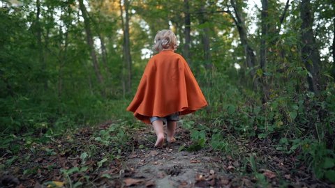 Little toddler boy cosplay gnome or hobbit in long cape walking barefoot in green forest. Halloween, kids concept. Amazing fairy tale character. Slow motion. 