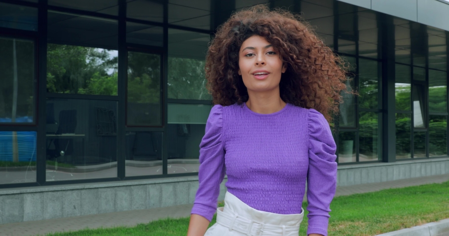 happy woman smiling and adjusting curly hair near building