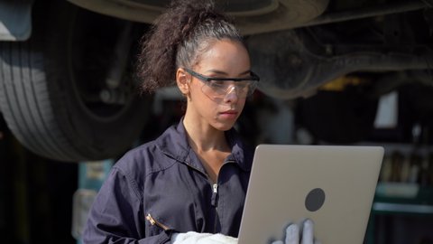 african american mechanic woman using laptop computer  checking list to undercarriage of car in workshop at auto car repair service center with lift . car engineer female inspection vehicle details
