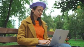 freelancer in autumn outfit typing on laptop on bench in park