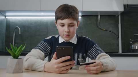 12-13 years old boy sitting at the kitchen table and shopping online with credit card and smartphone while sitting at home. Kid schoolboy with mobile phone using online payment systems. Online banking