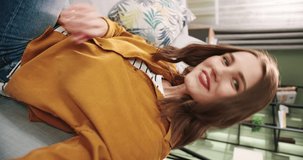 POV of Caucasian joyful smiling beautiful young woman sitting on sofa in cozy room recording video for blog. Female talks on video call through webcam at home and waving hand Close up Vertical concept