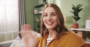 POV of Caucasian joyful smiling beautiful young woman sitting on sofa in cozy room recording video for vlog for social network. Video chatting on webcam at home and waving hand. Close up concept