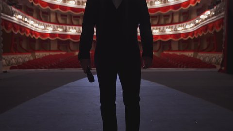 Man with microphone on theatre stage. Male actor in elegant suit walks on scene and starts speech in front of the empty auditorium. Back view.