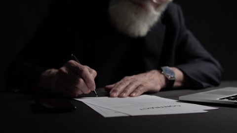Risky elderly boss with grey beard signing contract at table, business decision
