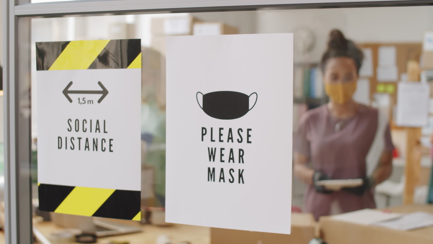 Close up arc shot of two paper posters with Social Distance and Please wear mask signs on glass wall in office on online shop; female colleagues in mask and gloves working in background Royalty-Free Stock Footage #1066214401
