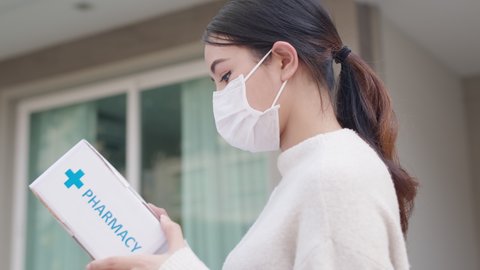 Young attractive beautiful asian female receive medication package box free first aid from pharmacy hospital delivery service at home wear glove, face mask in telehealth, telemedicine online concept.