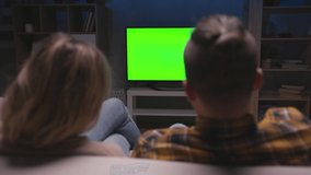 Family Couple Watches Green Screen TV Mock-up Sitting on Couch in Living Room Together. Rear View on Casual People who Watching TV Green Screen in Domestic Cinema. Looking TV Show or News in Home Rest