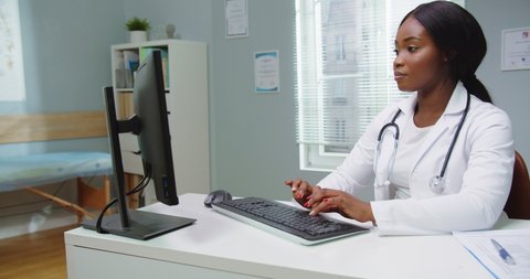 Medium shot of beautiful young african american woman doctor assistant working in medical office using computer, looking in camera smiling. Modern healthcare, online medical help concept