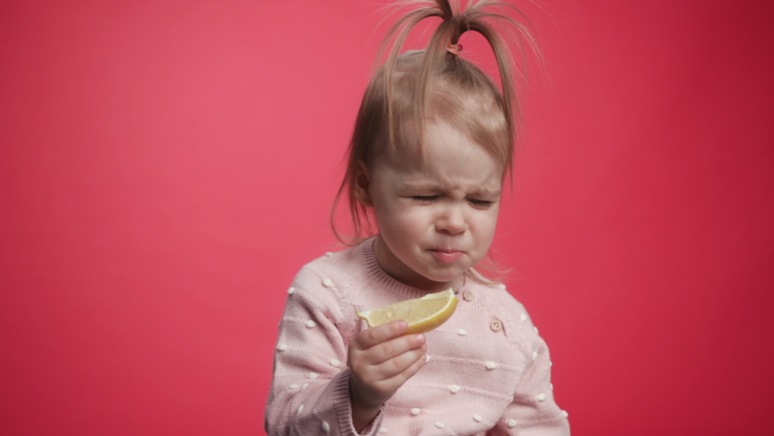 One Cute Beautiful Baby Eating a Lemon. A Child Tastes the Raw Lemon. Tiny Playful Girl Eats Fruits. Young Kid Holding Eat Tasty. Fun Expression Emotion Faces Kids from Eating Sour Lemon. Bright Look