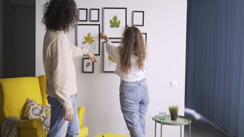 Romantic couple in jeans and pullovers hangs different sized wooden frames with coloured dried leaves on wall decorating new flat backside view