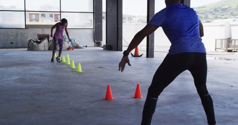 African american woman doing slalom with a football in urban building with man cheering her. urban fitness and healthy lifestyle.