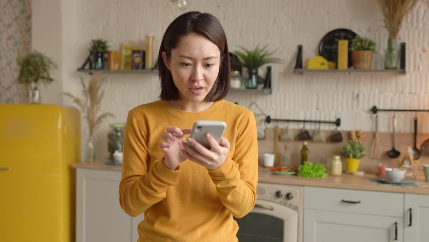 Excited mixed race female customer celebrating getting ecommerce shopping sale offer on smartphone at home. Asian woman winner looking at mobile phone using app celebrating success concept Royalty-Free Stock Footage #1066234381