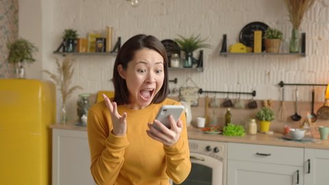 Excited mixed race female customer celebrating getting ecommerce shopping sale offer on smartphone at home. Asian woman winner looking at mobile phone using app celebrating success concept