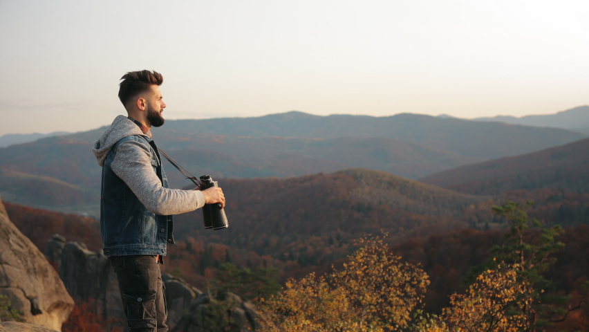 The man is standing on the edge of a cliff and looks through binoculars. Mountain landscape in the background. Hiking in the mountains. 4K.