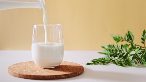 Pouring vegan milk in glass from a bottle on a light neutral background with space for text, copy space. Glass of vegetable milk drink for healthy vegetarian in slow motion. 