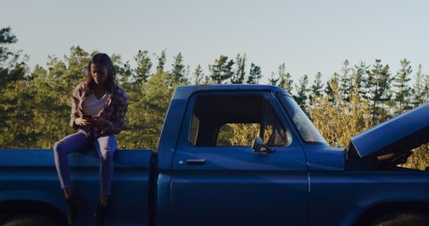 Front view of a young mixed race couple by a broken down pick-up truck, the woman sitting on the car using a smartphone, the man standing by opened hood during a road trip