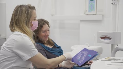 Female orthodontist showing a patient a dental pamphlet or brochure in a clinic. Woman looking at booklet explaining about medical procedure for teeth alignment using invisible retainer or Invisalign