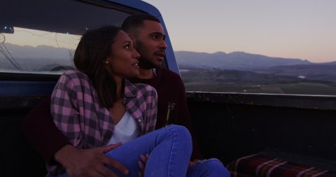 Front view of a young mixed race couple sitting on the back of their pick-up truck, drinking beer, the man embracing the woman during a road trip