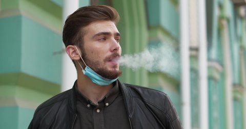 Young handsome man in medical protective face mask on the chin smokes a cigarette, then he takes off mask. Cinema 4K 60fps video