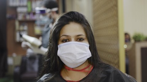 front view close up shot of a young attractive Indian woman with long black hair sitting in a salon with spa apron and protective face mask on looking at the camera 