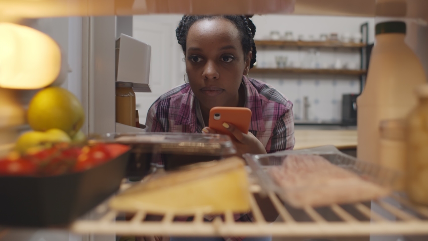Afro-american woman looking inside refrigerator making grocery shopping list on mobile phone app. View from inside fridge of african female ordering food delivery on smartphone Royalty-Free Stock Footage #1066262053