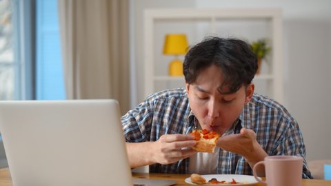 Young asian man biting slice of pizza when reading e-mail from colleague on laptop screen. Portrait of korean student eating pizza for dinner working on project using computer at home