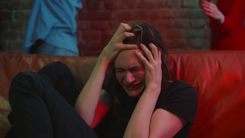 Young man crying hallucinating after drug overdose at club party. Close up portrait of drug addict guy having hysteria sitting on couch with people dancing on background in nightclub