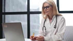 Female MD wearing white coat and stethoscope, communication with a patient via video calling on laptop. A doctor is listening complaints and takes notes conducting virtual consultation. Telemedicine
