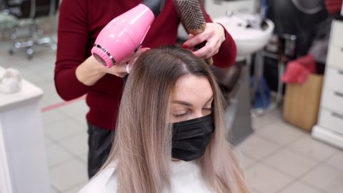 a hairdresser combs her hair with a round brush and dries it with a hairdryer to a girl in a protective mask