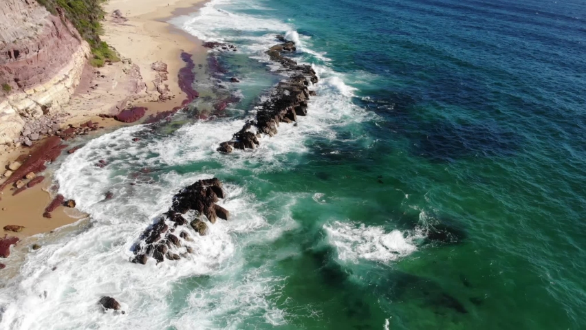 Flying along Pembula Pinnacles and the Merimbula Headland, South Coast of NSW, Australia. Waves crashing over red, orange and yellow rocks, aerial views, bushland meets the sea. Holiday here this year Royalty-Free Stock Footage #1066272397