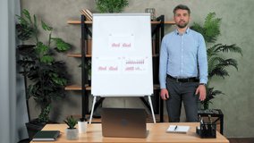 Smiling man teacher tutor stands near flip chart greets waves hand listens student question remote video call chat. Coach trainer records online business webinar master class course in home office