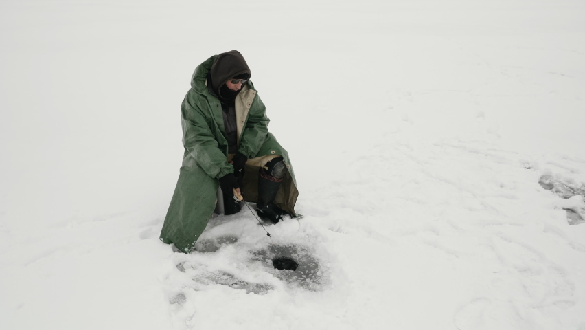 Adult male is fishing in ice hole at winter fishing. Fisherman is catching fish on frozen lake. Man with fishing rod is sitting on ice pond