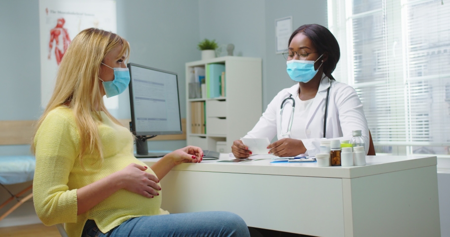 Medium shot of pregnant woman with medical mask visit african american gynecologist at hospital. Doctor in glasses giving ultrasound picture. Pregnancy woman and maternity healthcare concept. | Shutterstock HD Video #1066286389
